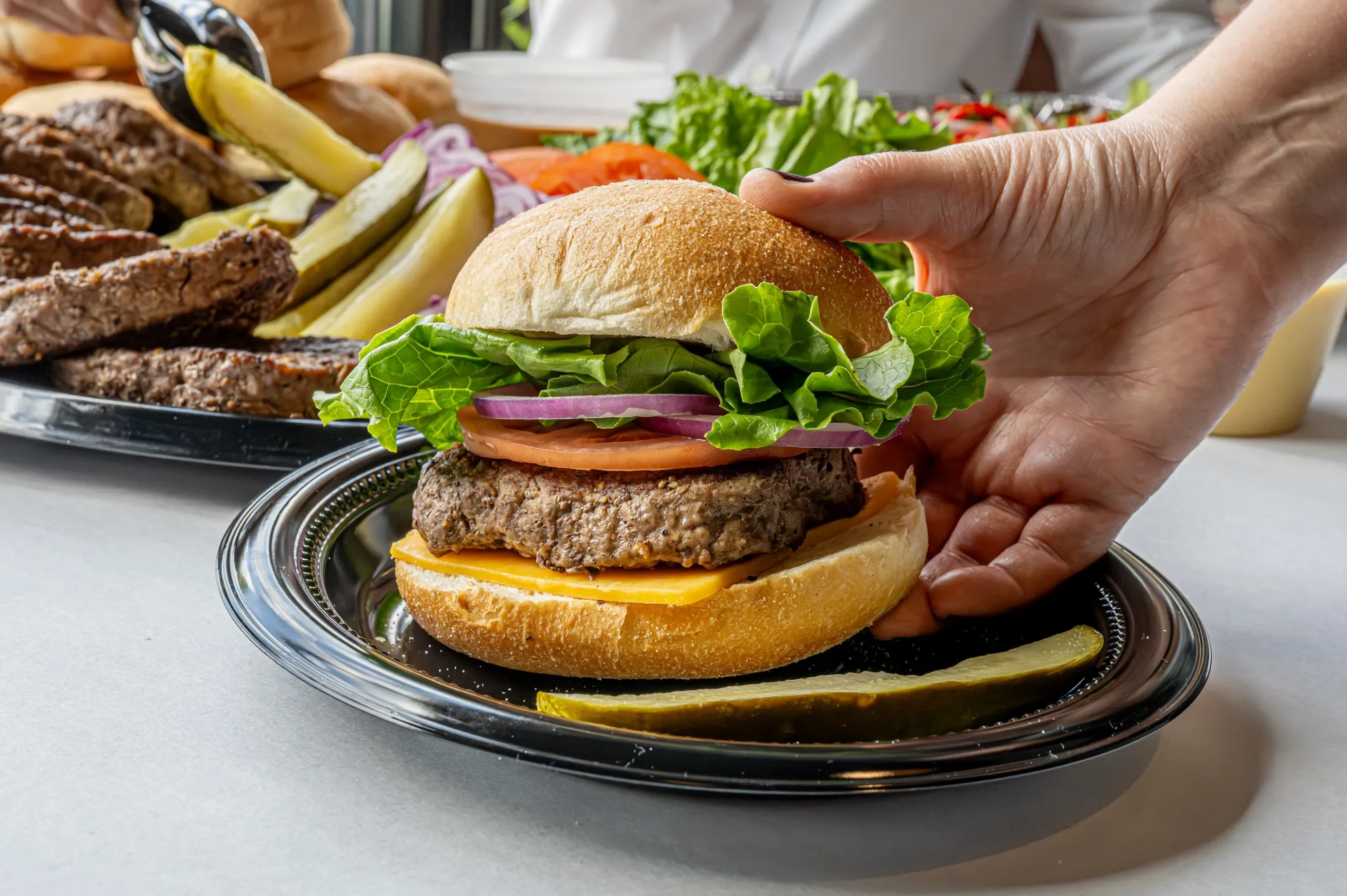 A cheeseburger being picked up with patties and pickles in the background